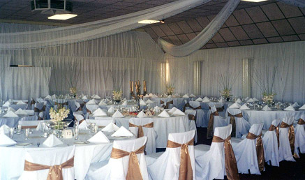 Large function room set up for a wedding reception with white drapery on walls, white tablecoths on tables, white chair covers and floral arrangements on tables